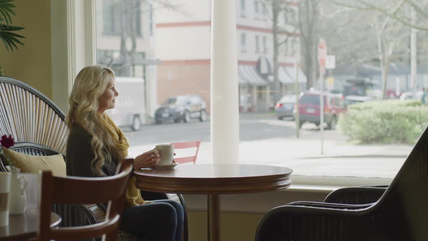 Blonde woman holding the coffee mug sits at the tableand looks out the window in a cafe during daylight