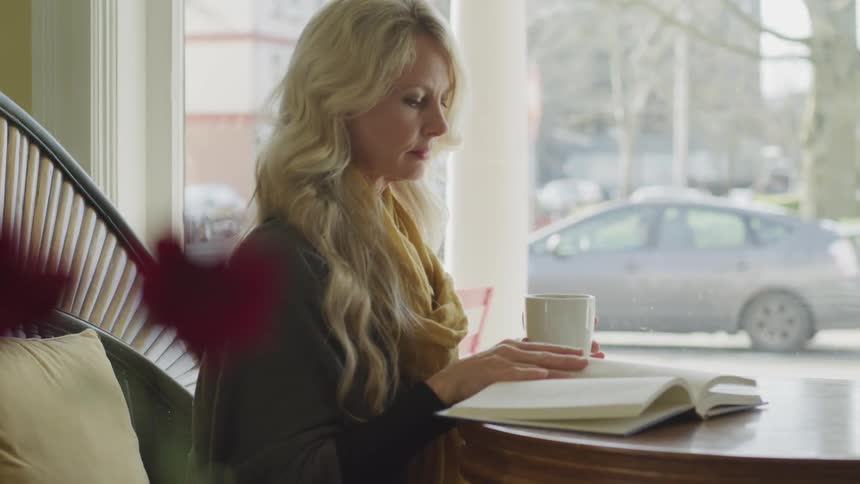 Blonde woman sittingat the table reads a book and takes a sip of her coffee in a cafe