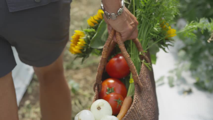 A woman walks and carries a straw bag filled with flowers tomatoes and onions