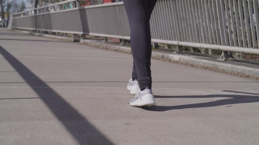 Someone in dark blue leggings and gray sneakers walking down a sidewalk beside a metal fence