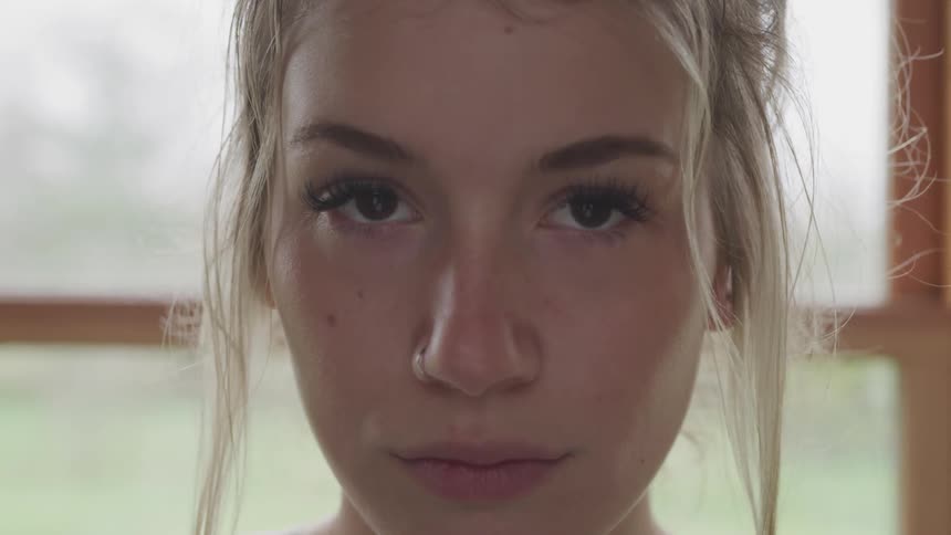 A blonde woman with a nose ring stands in front of a paned window