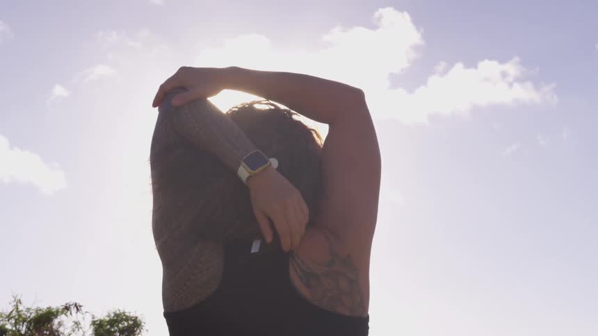 A tattooed woman doing shoulder stretches outdoors during the day