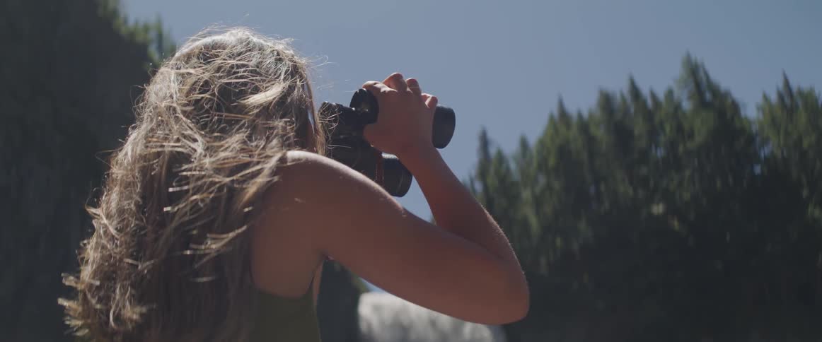 A woman holds binoculars in her hands and gazes into some evergreen trees