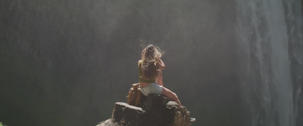 A young woman with long brown hair sits atop a tree stump near the base of a small waterfall