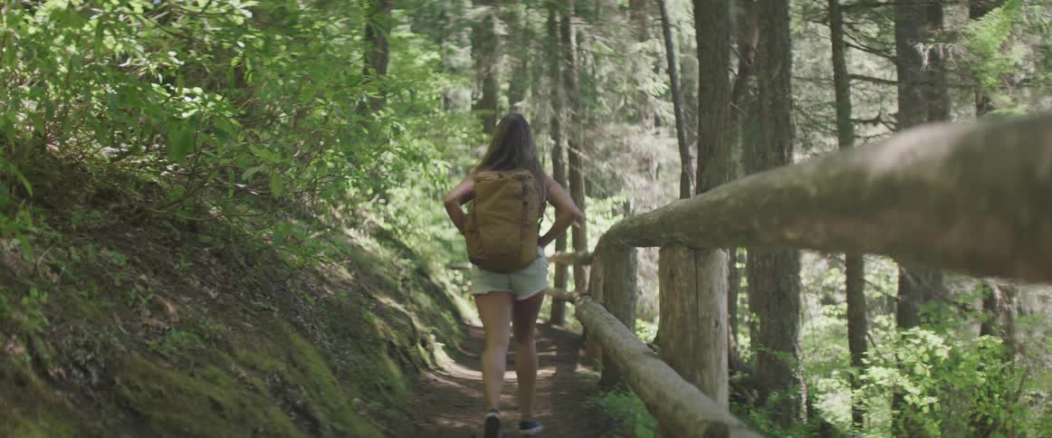 Girl wearing white shorts and carrying brown backpack walks along a fenced path on a forested hillside