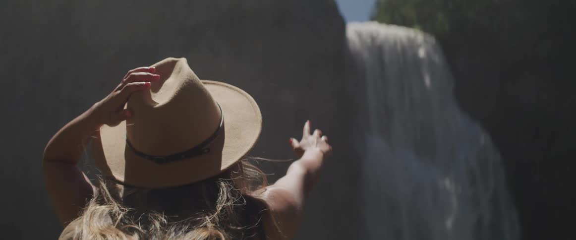 A woman holds on to her hat as she reaches her hand out towards a waterfall and the water mist