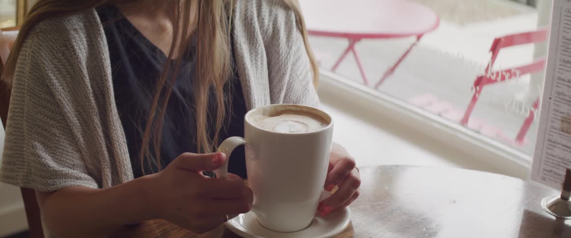 A woman sits at a table in a cafe by the window with a large mug
