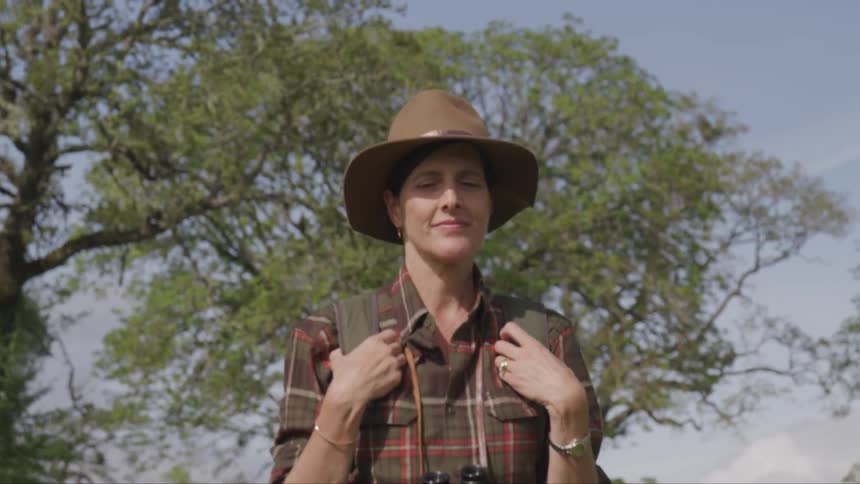 A white woman wearing a brown hat walks through a forested area
