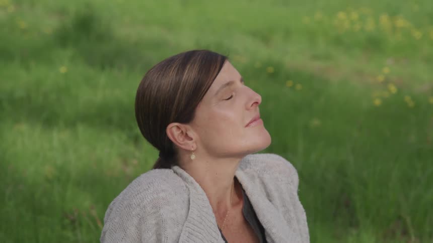 A woman sits in a field with her eyes closed and her head up towards the sky