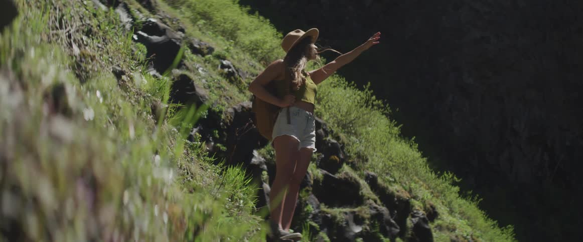 A female in a widebrimmed hat and carrying a backpack stands on the side of a mountain reaching towards something in the sky