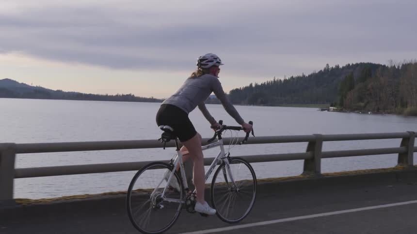 A tall woman pedals her bicycle along a road that borders a large body of water that is surrounded by forested hills