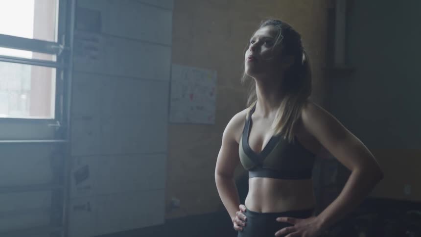 A woman in a black sports bra and leggings breathes heavily while standing in a gym and looks up and down