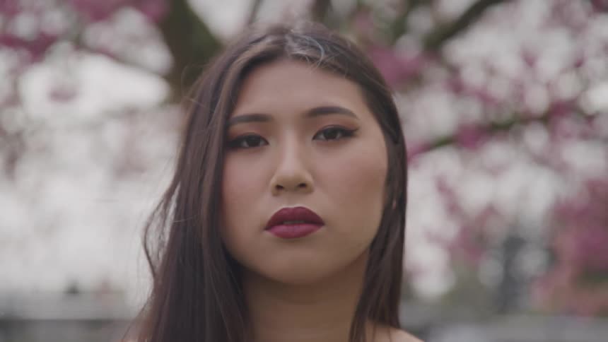 Asian woman looks at camera with lips matching the color of leaves on the tree behind on a cloudy day