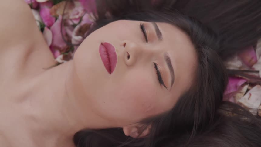 A female lay on the ground with her eyes closed surrounded by flower petals