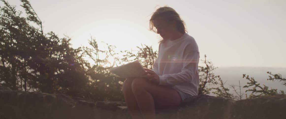 Woman in white shirt and shorts sits on rock wall at edge of valley holding paper