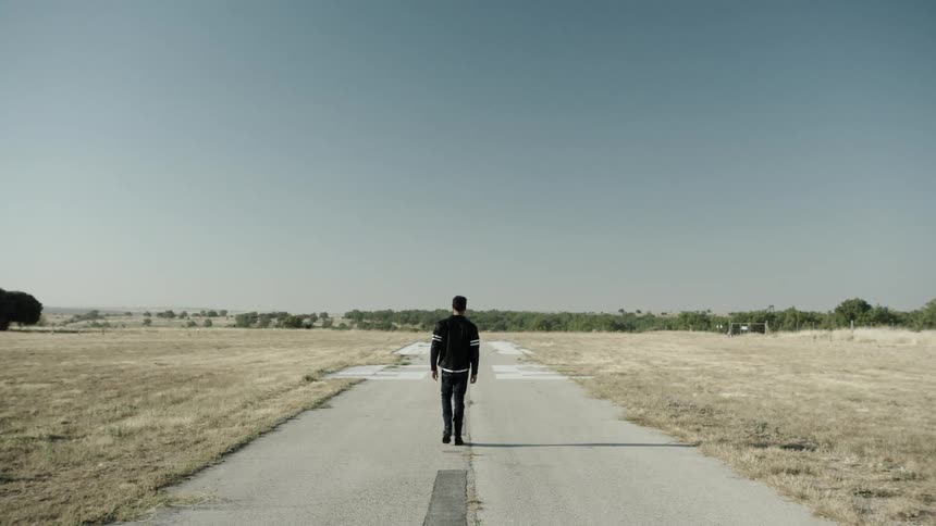 A man walks on an empty and cracked strip of pavement in a field on a sunny day