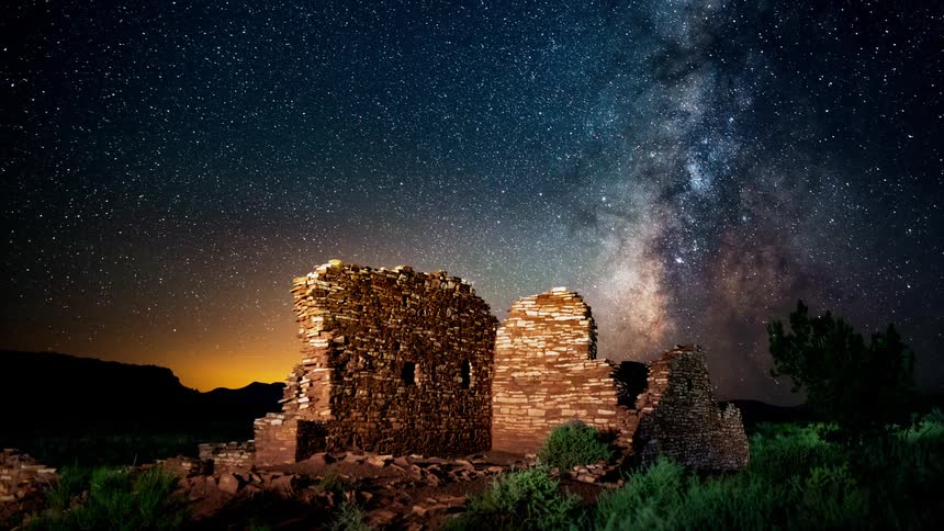 The Milky Way moves across the sky behind a lit rock formation