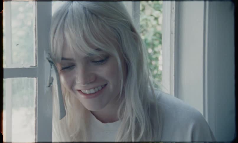A blonde female sits on a window sill smiles and then looks at the camera