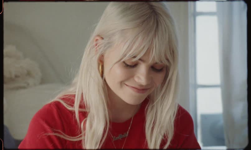 A blonde haired pendant wearing woman sits on a bed and reads a hand written note