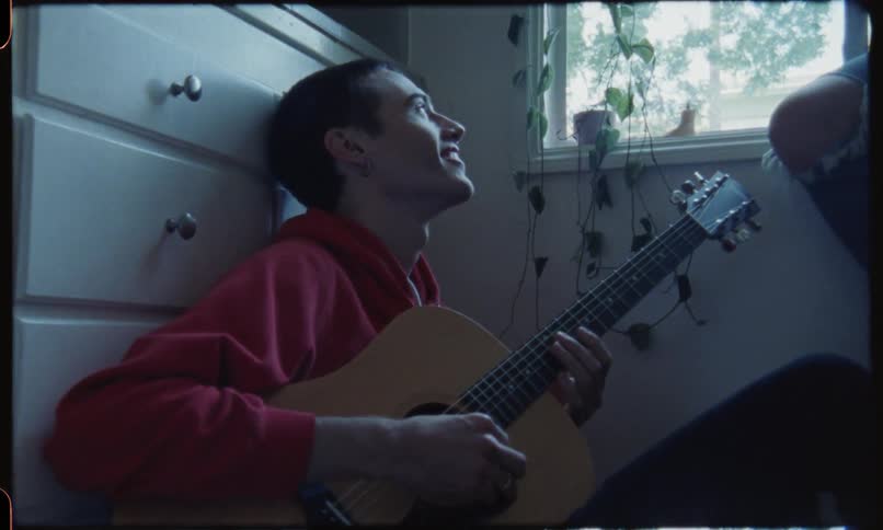 A man plays guitar on the floor while a woman sitting on a windowsill watches