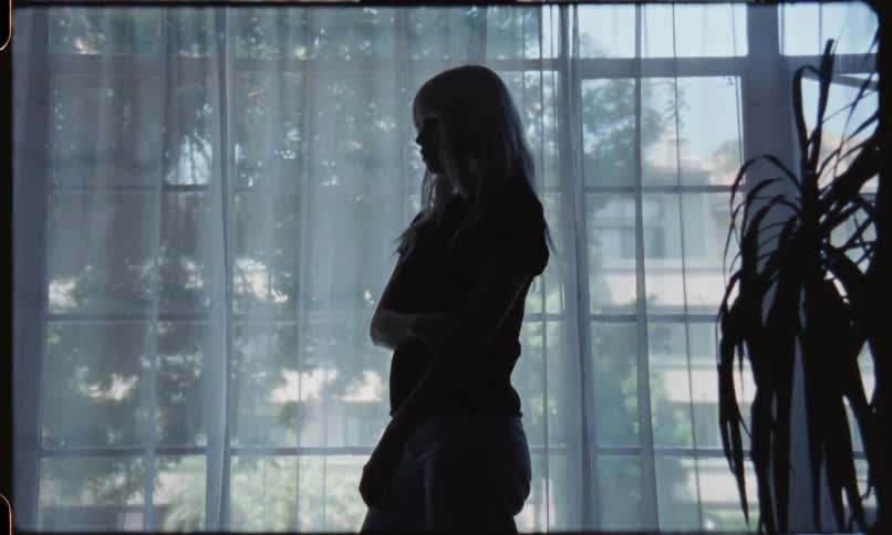 A female with blonde hair is standing in front of a wall of windows with one arm across her body holding onto the elbow of her other arm