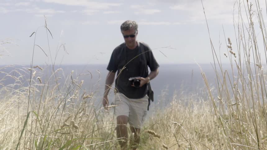 A man in shorts carrying a camera walks in a grassy field high above the ocean seen in the background