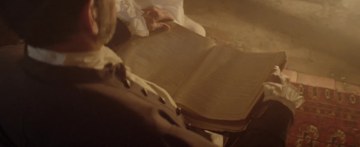 A man in civil war era garb holds a book in his hands and reads aloud by fire light