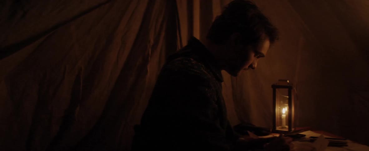 A man studies by candle light at a desk inside of a tent
