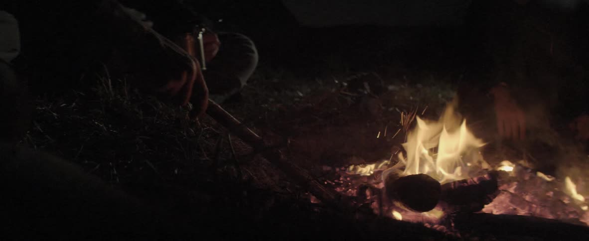 A fire in an open pit is tended by someone holding a long stick