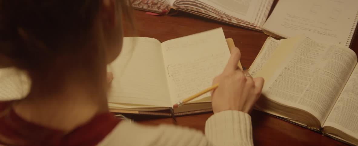 A young woman writes in a book while other books are spread across the desk