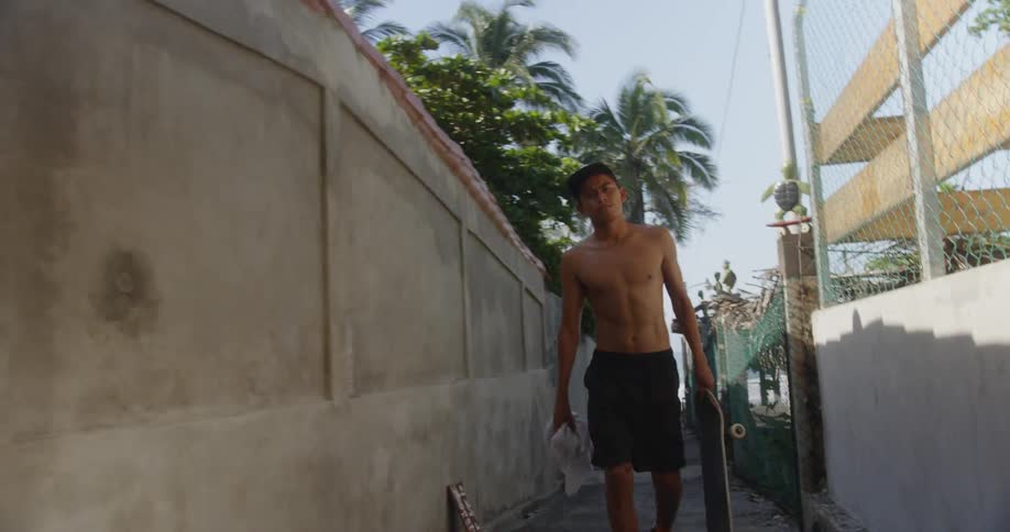 A shirtless hispanic teenager walks down an alley while carrying a shirt and a skateboard