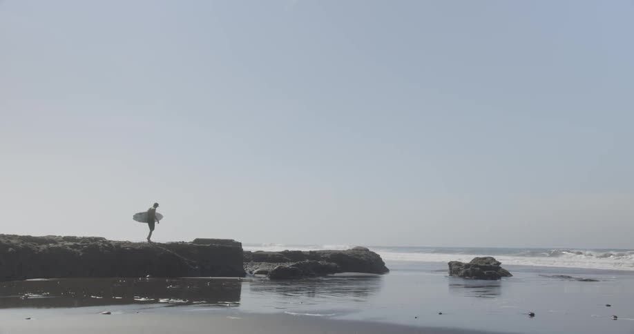 Man carrying surfboard walks toward ledge overlooking beach
