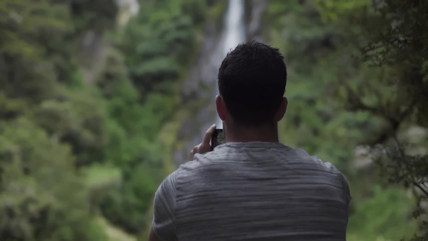 A man takes a photo of a waterfall cascading in a natural setting