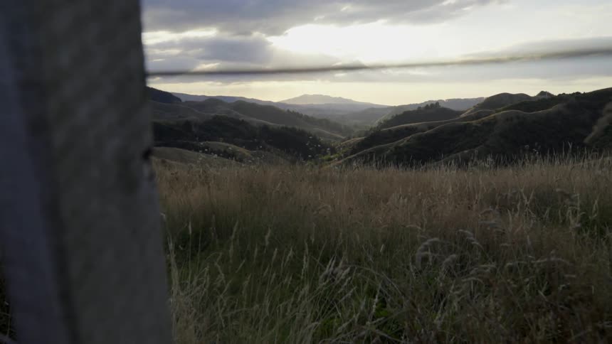 A field of grass with hills in the distance is seen through a wood and wire fence