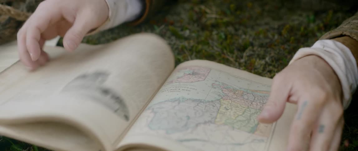 A man reads an old geography book while wearing period clothing
