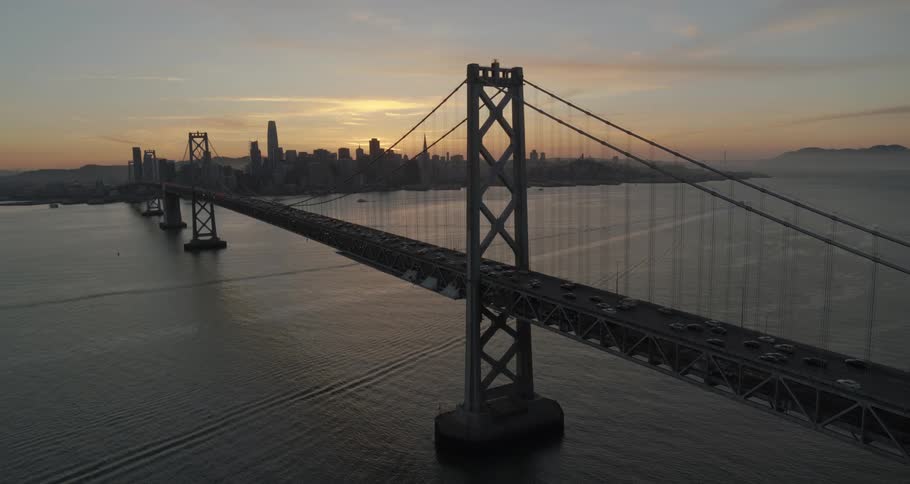 Traffic crosses suspension bridge as fog covers ocean near downtown city as sun rises