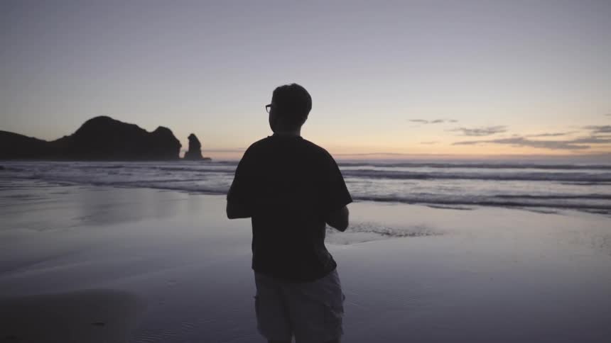 Man Standing on Sandy Beach at Sunset Watching Ocean Under Warm Evening Sky.