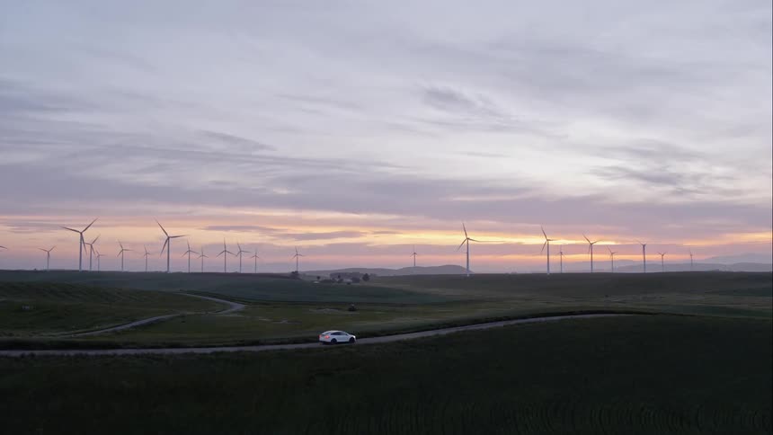 A white car drives a long an isolated road next to a wide open field lined with multiple windmills