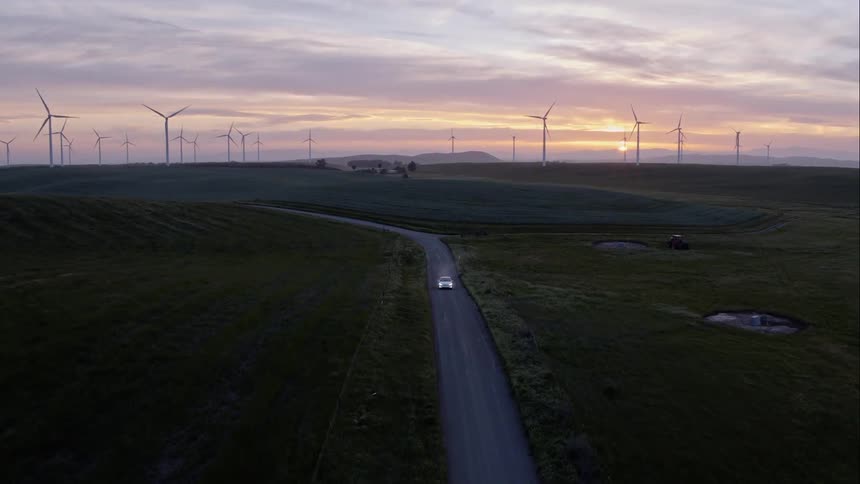 A car with headlights on travels on a dirt road through fields with wind turbines on the horizon