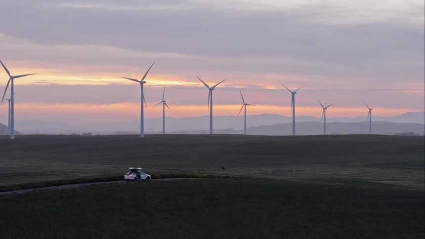 A white car opening its doors and a person coming out as wind turbines rotate in the background