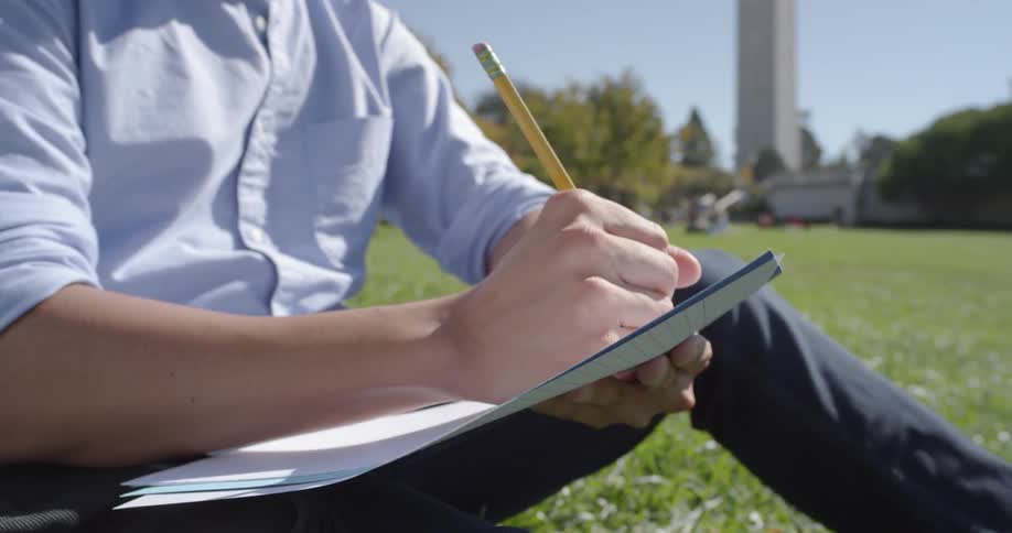 Man writes on a large paper as he sits on the grass under a sunny sky