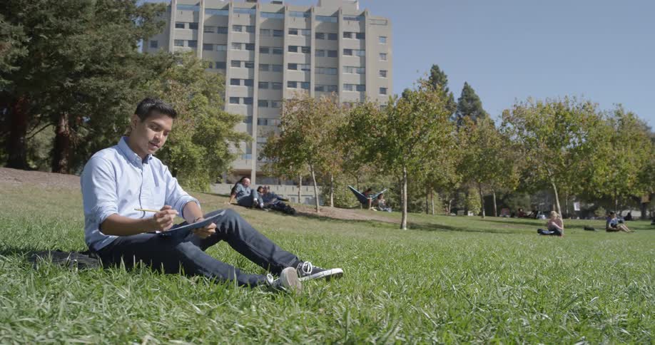 A man sits on the grass in a park and writes resting papers on his knee
