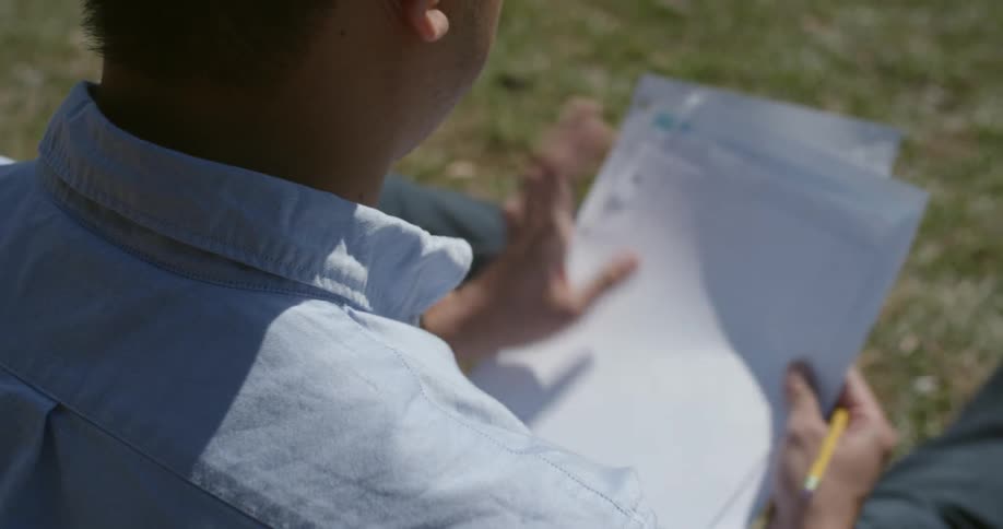 A man sits on the grass and shuffles papers until he begins to write