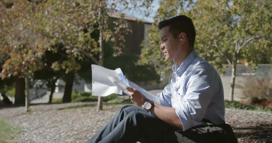 A young man is reading some papers in the shade of a tree while sitting on the ground