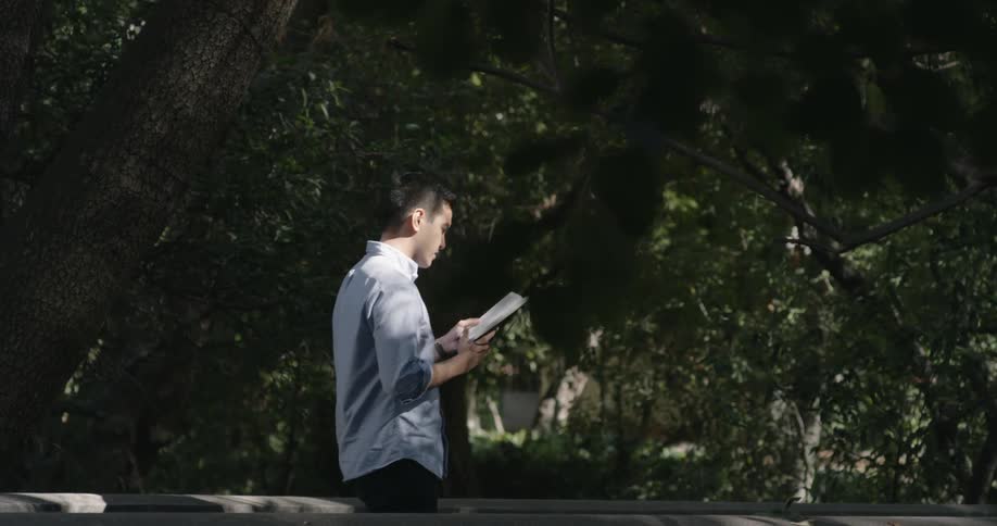 Man walks back and forth near the trees as he reads a book
