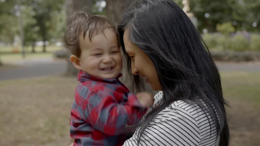 Woman cuddles toddler boy and laugh together in the public park during daylight