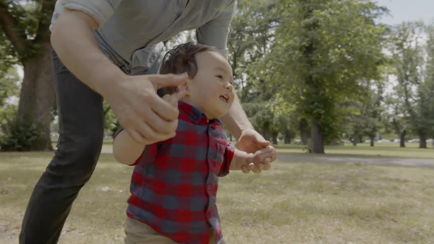 Man helps toddler boy walk on the grass in the public park during daylight