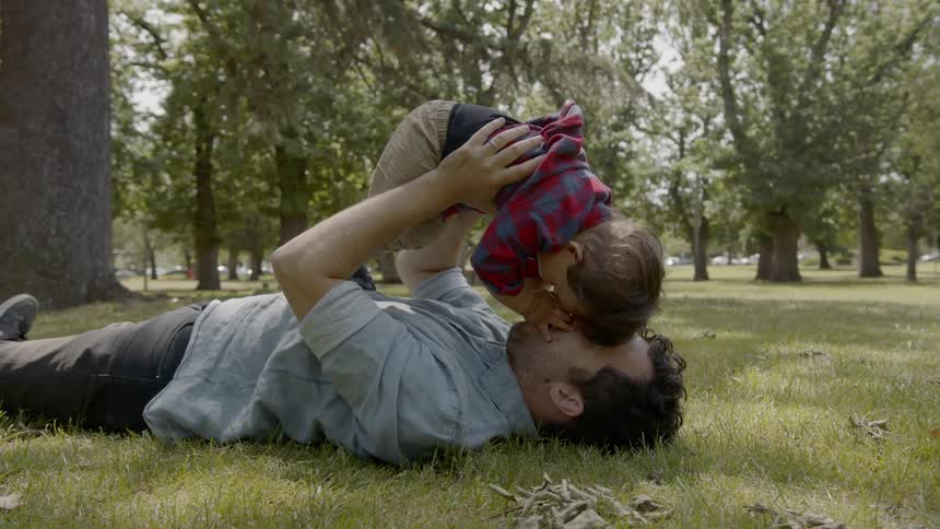 A man lies in the grass with a little boy in his arms that he is lifting above his chest