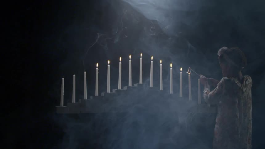 A young black female is carefully extinguishing a one of several candles in a darkly lit room that is filled with smoke