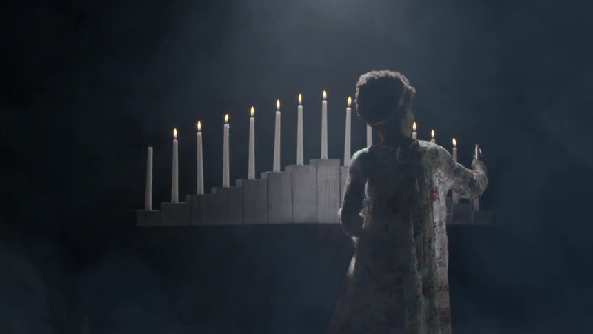 A woman stands before a row of candles and she uses a long candle snuffer to extinguish one on the end of the row